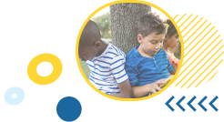 2 young children, a black boy and a white boy sitting leaning against a tree while reading.